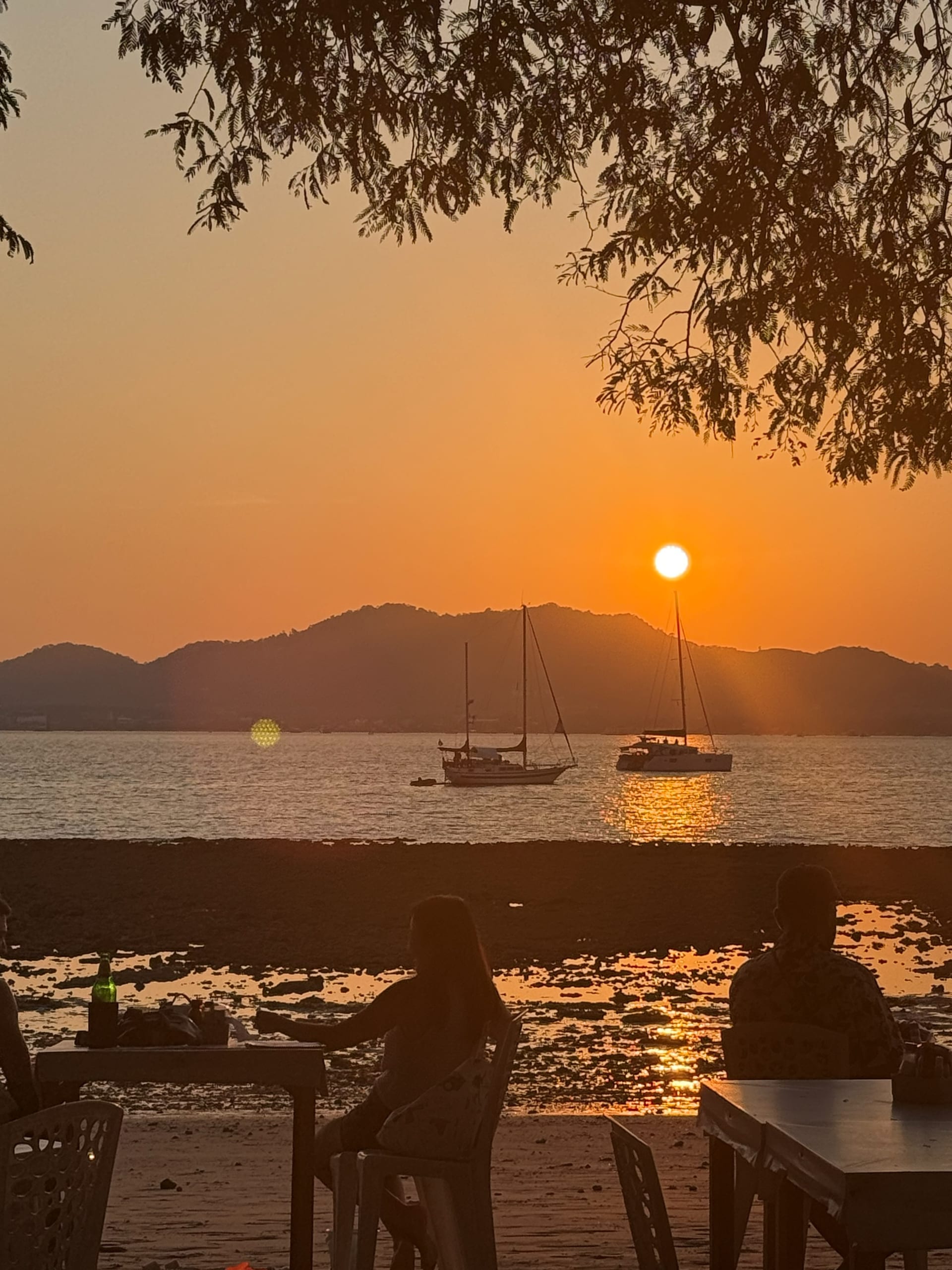 At The Beach Restaurant & Bar earlier in the evening — sun still above the mountains, warm golden wash over Ao Yon Bay, two diners silhouetted with plastic chairs at the water's edge, two sailboats moored, exposed beach floor at low tide
