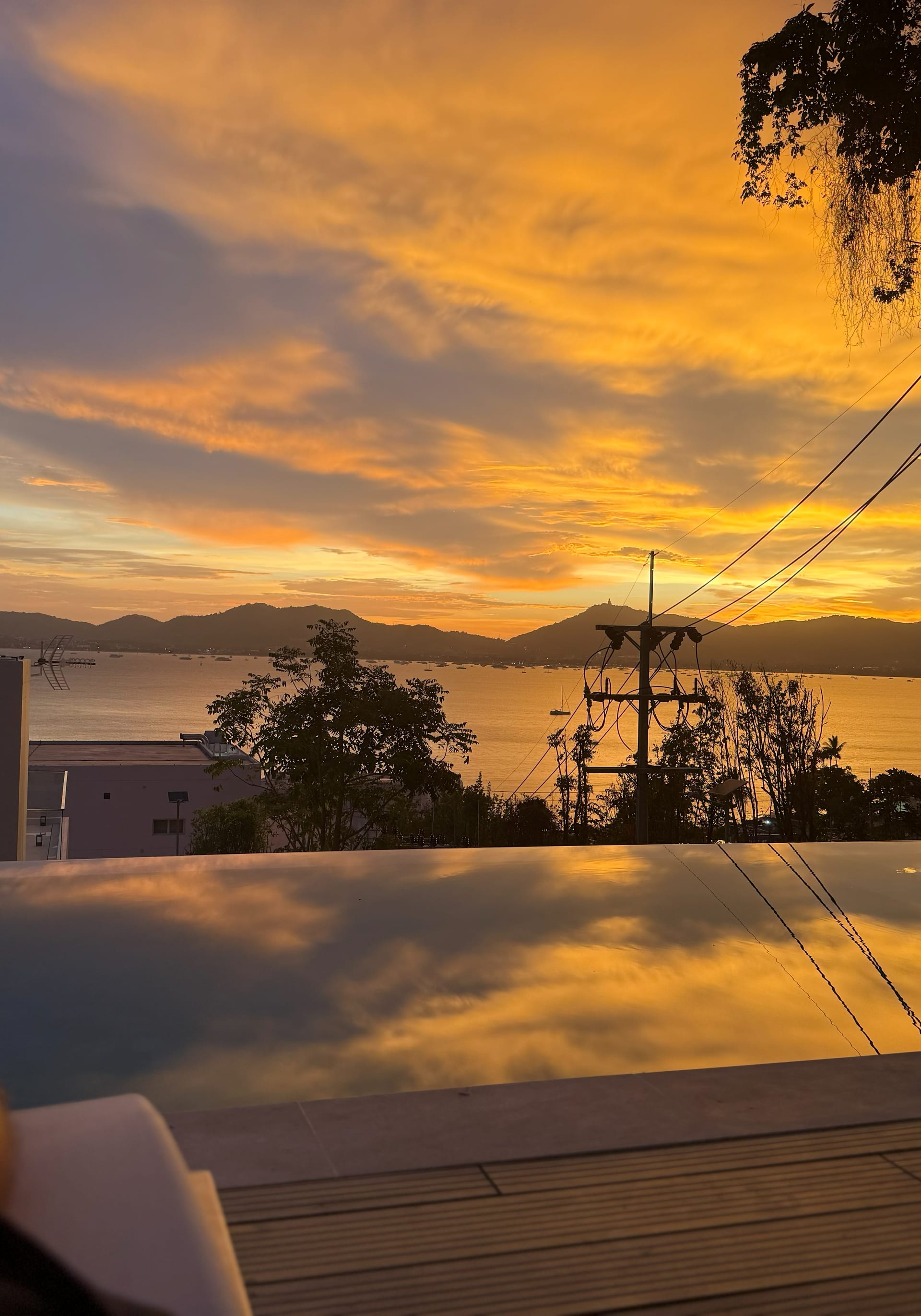 Fiery orange and gold sunset over Ao Yon Bay from At The Beach Restaurant — dramatic cloud structure mirrored in the infinity-pool deck, palm-covered mountains silhouetted, neighbourhood power lines crossing the right side as part of the authentic Cape Panwa setting