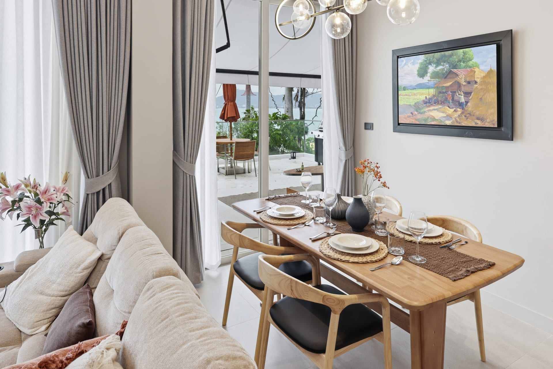 The wood dining table set for four beside the linen sofa and framed art, sea view beyond