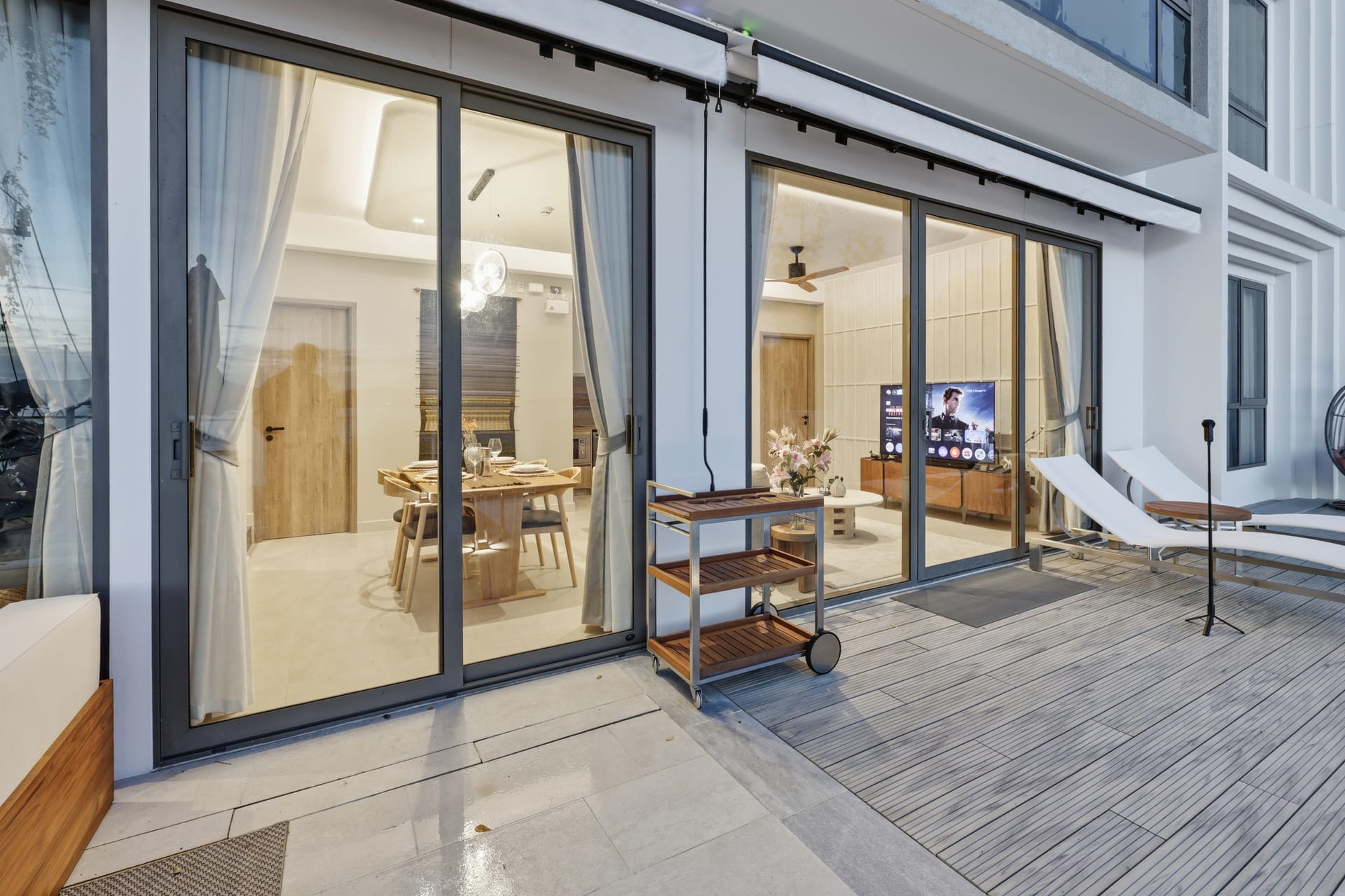 The white villa at dusk — full-height great-room glass glowing warm, the pool terrace and bay beyond