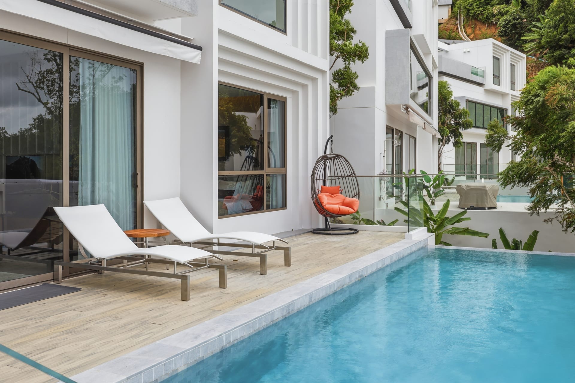 The villa exterior in daylight with the infinity pool, sun loungers, a hanging chair and tropical greenery