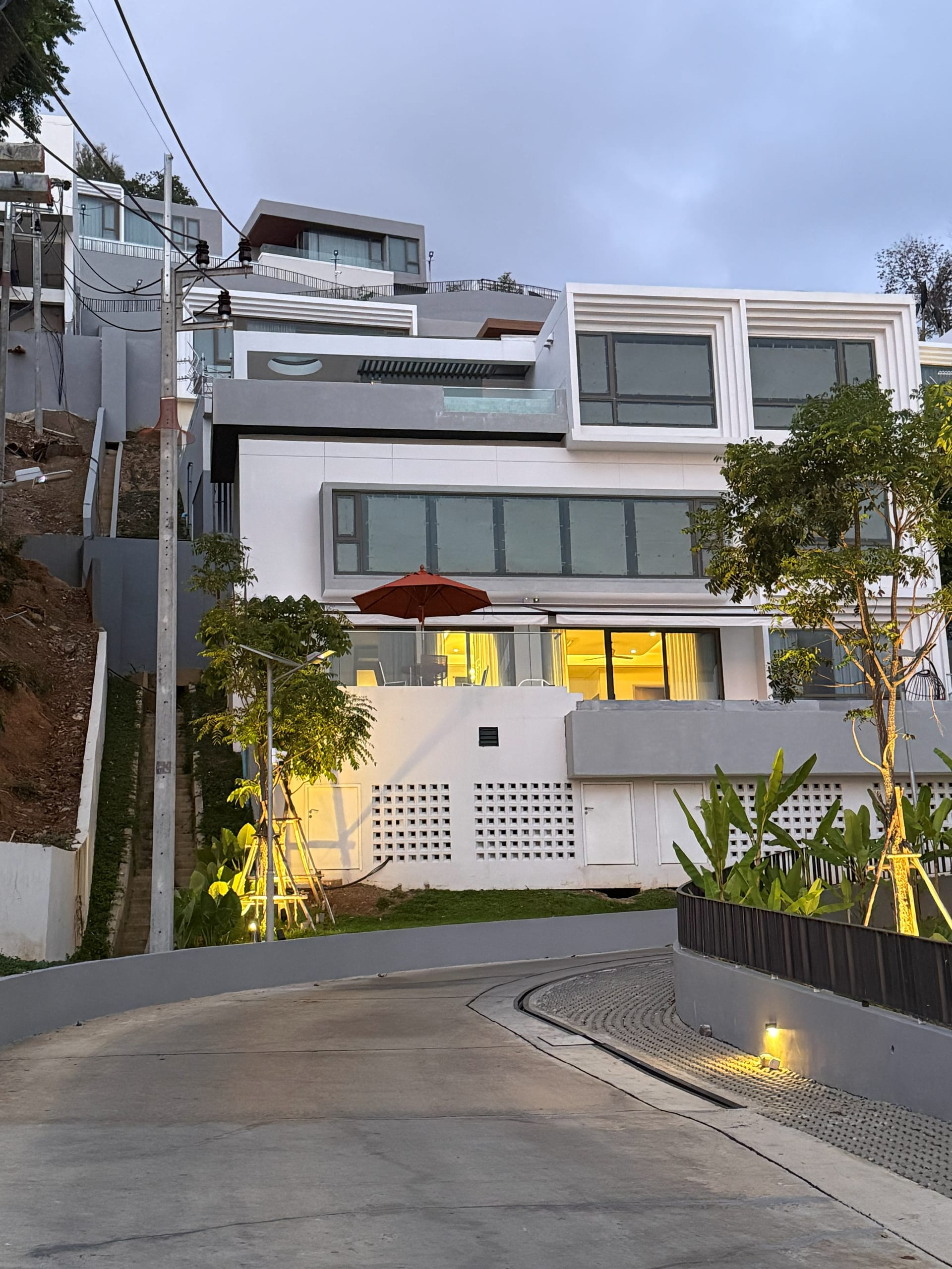 Three-storey white villa at dusk photographed from the driveway approach — warm interior light glowing through the great-room glass, red parasol on the upper terrace, banana plants and tropical greenery framing the lower entrance, hillside behind