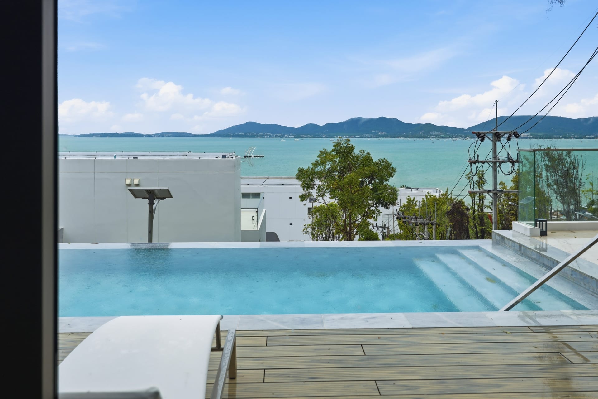 The private infinity pool on a clear day — timber deck and infinity edge overlooking Ao Yon Bay, green headlands and the Andaman under a bright blue sky
