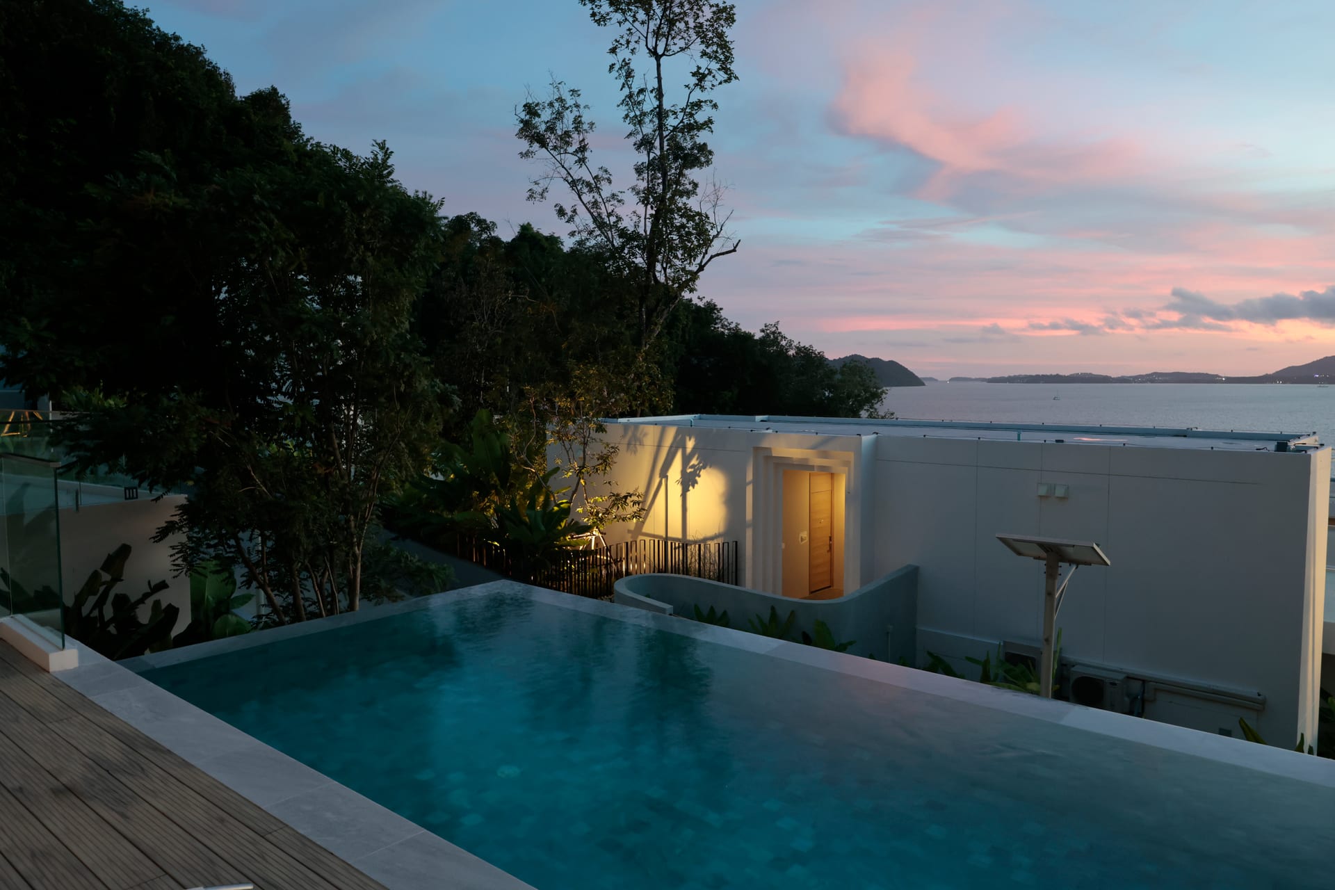 The infinity pool at dusk — soft pink sky, the villa's warm-lit doorway and the bay beyond the deck