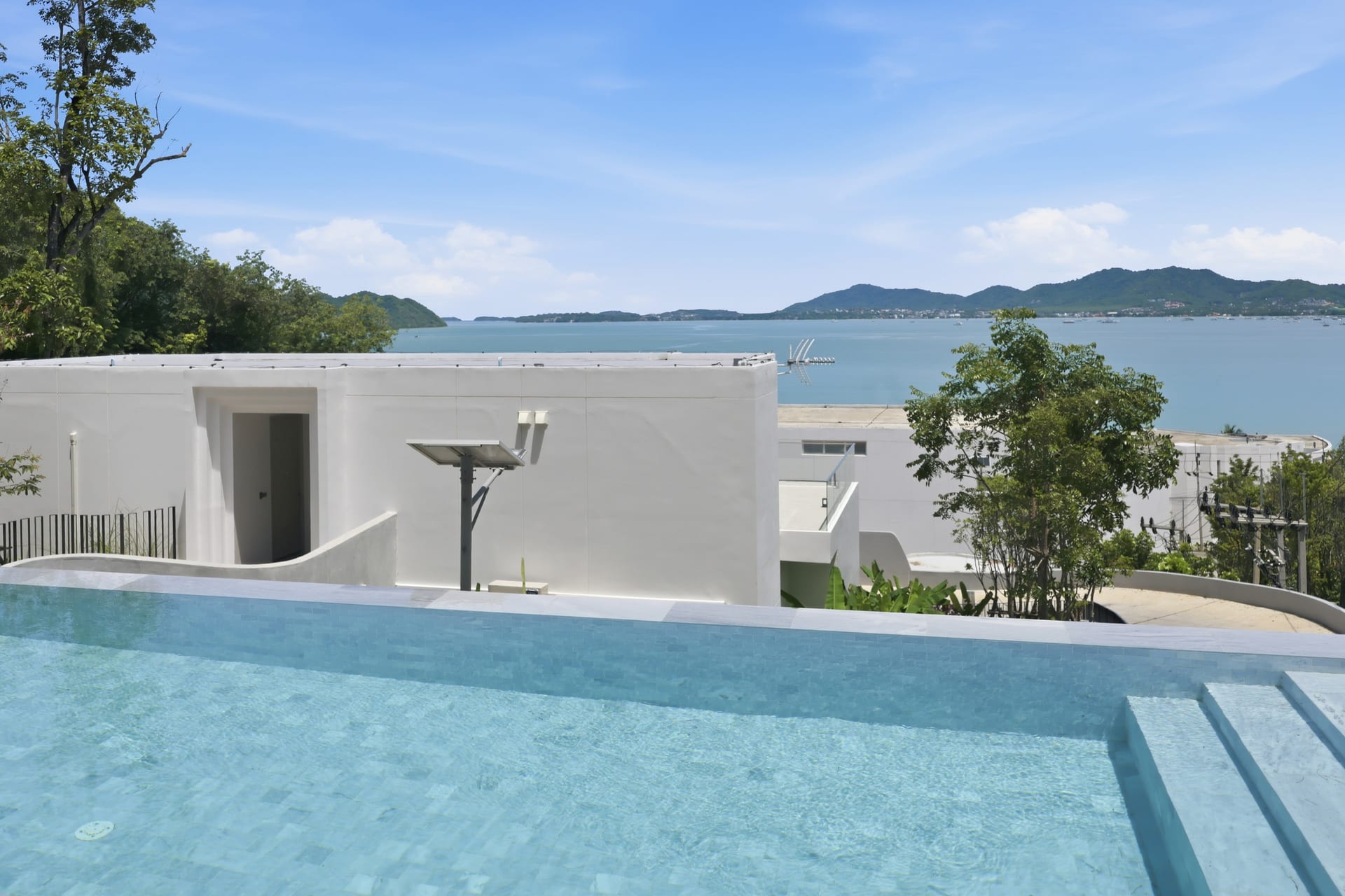 Wide view of the infinity pool with sun loungers on the teak deck, the bay and Cape Panwa hills beyond