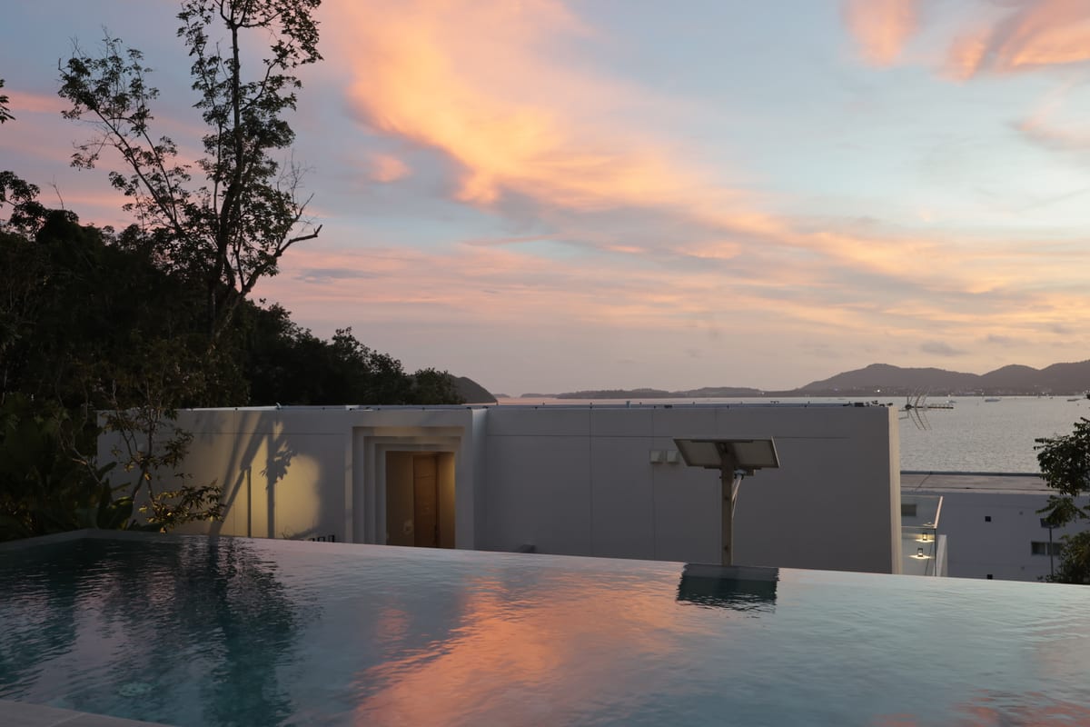 The infinity pool at sunset — a pink and orange sky reflected in the still water, the white villa and Ao Yon Bay beyond the timber deck