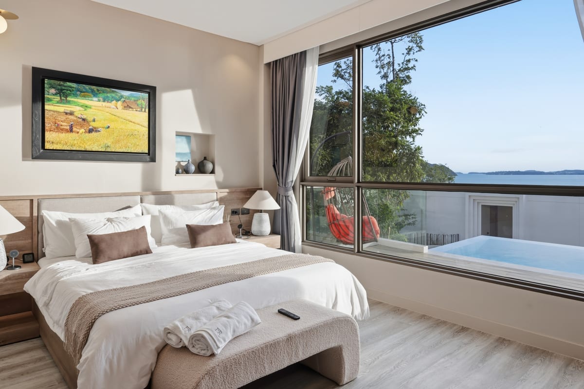 The primary bedroom's king bed facing floor-to-ceiling glass, the bay and Cape Panwa hills beyond the sliding doors