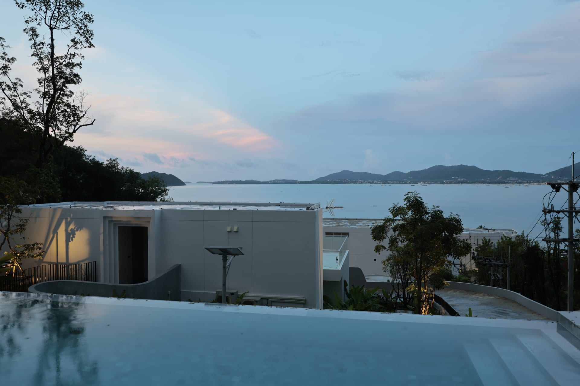 Sunrise over the suite's infinity pool and Ao Yon Bay — soft early light on the white villa, calm Andaman water and distant headlands beyond the timber deck