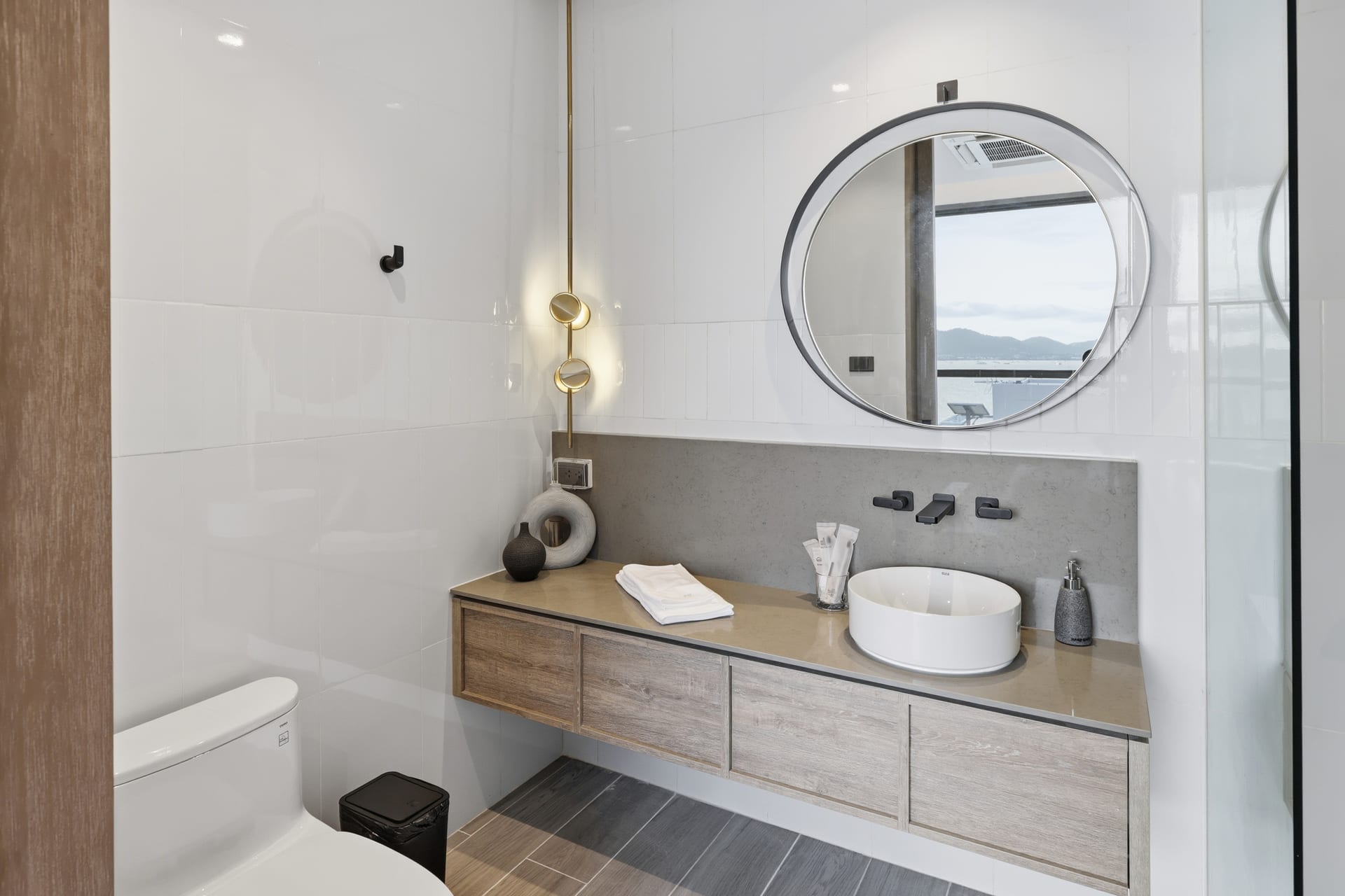 The primary bathroom vanity with a round mirror and a sea-facing window, toilet alongside
