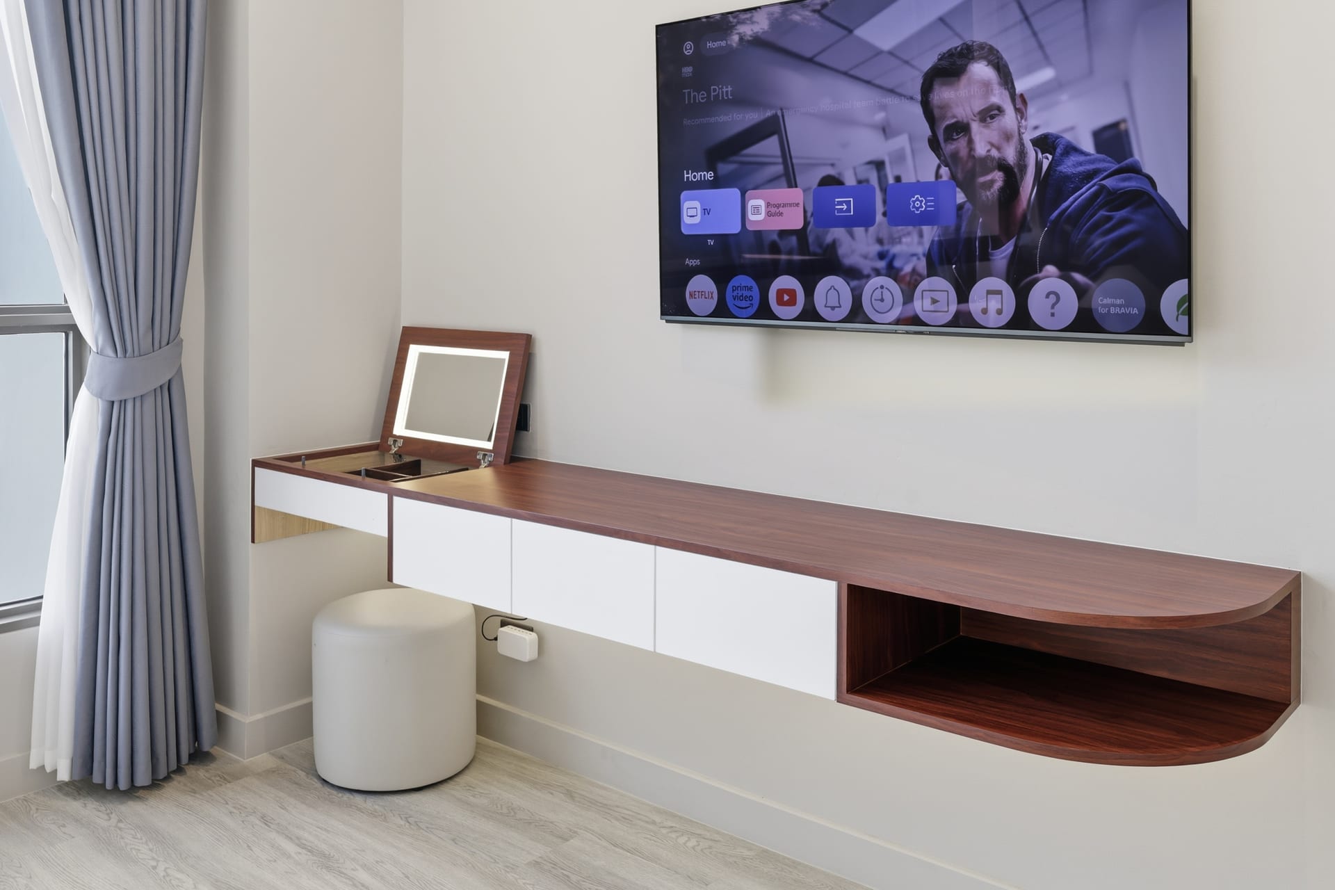 A floating desk and console with a wall-mounted TV and a stool, sheer curtains alongside