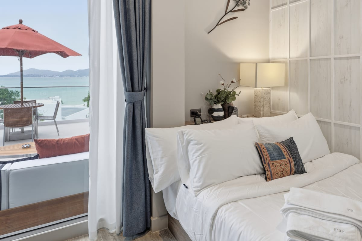 The second bedroom with a queen bed in white linen, sliding door to a private balcony with a red parasol and the bay beyond