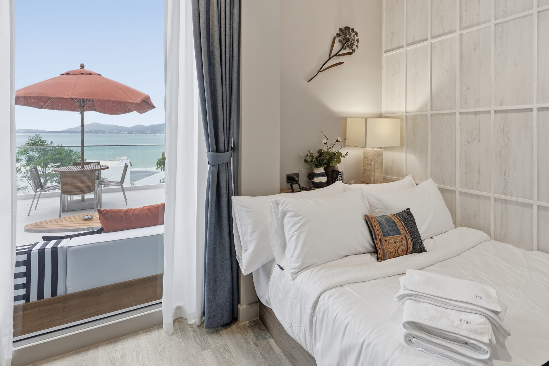 The second bedroom's queen bed and bedside lamp, sheer curtains framing the balcony door, a red parasol and the bay outside