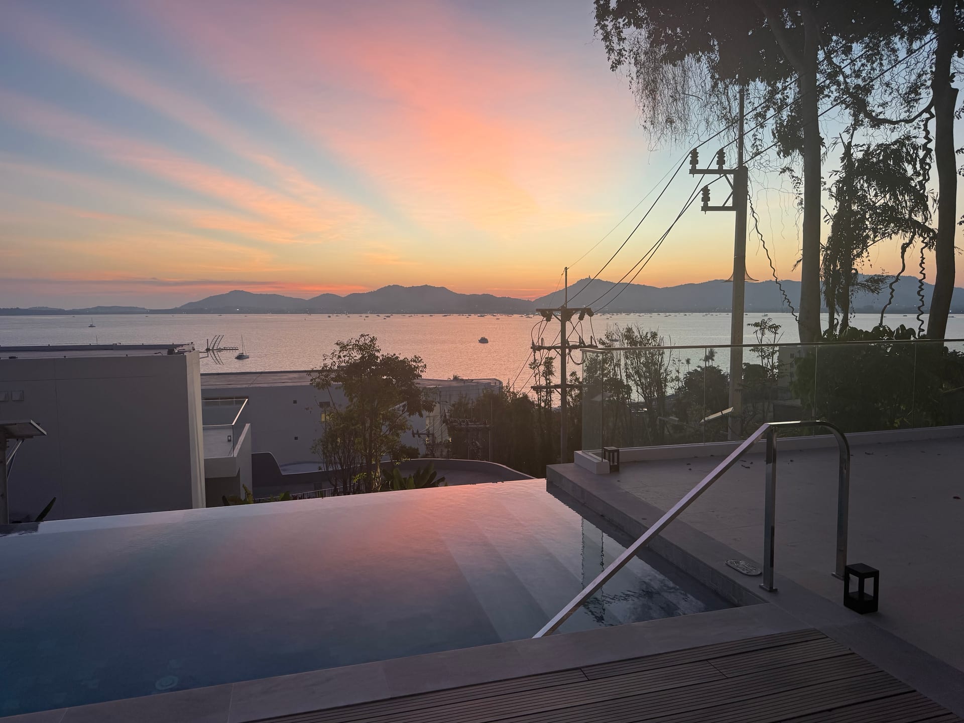 Sunset from the suite pool deck — pink and orange sky over Ao Yon Bay, Phuket mountains in the distance, the Andaman Sea below, longtail boats and yachts moored on the still water