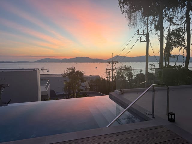 Sunset from the suite pool deck — pink and orange sky over Ao Yon Bay, Phuket mountains in the distance, the Andaman Sea below, longtail boats and yachts moored on the still water