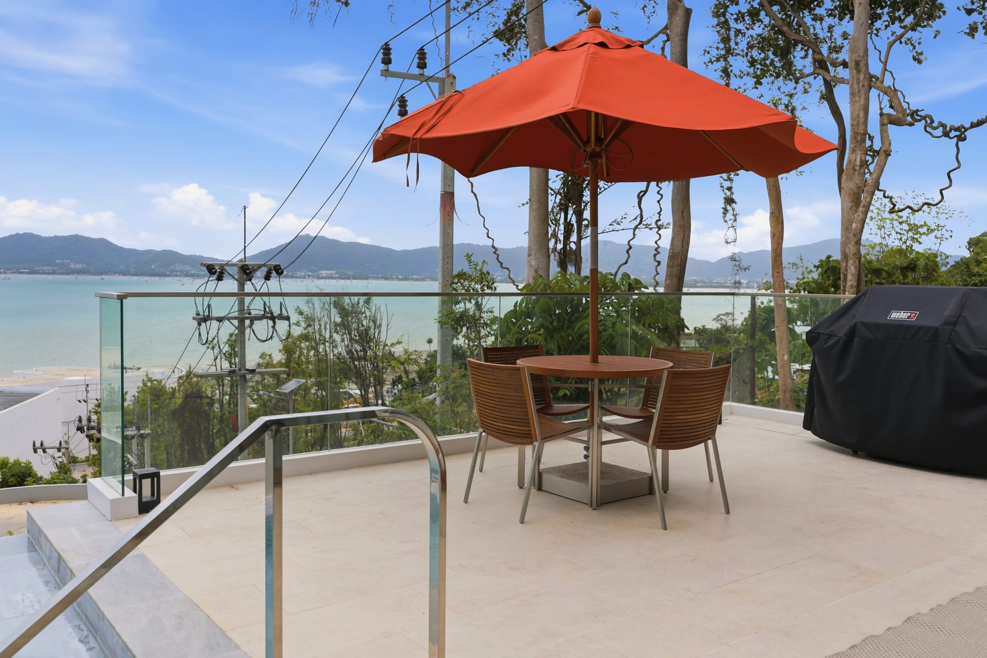 The pool terrace with an orange parasol, a daybed and an al-fresco dining set, the bay and hills beyond the glass railing