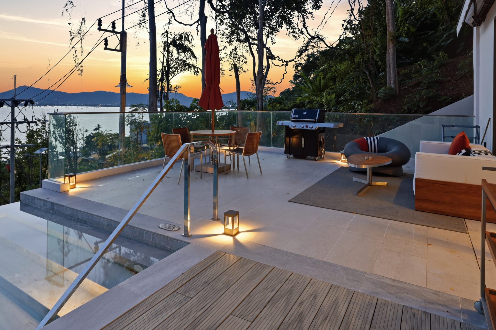 The terrace at twilight, lit lounge and dining areas under the parasol, the bay glowing in the last light