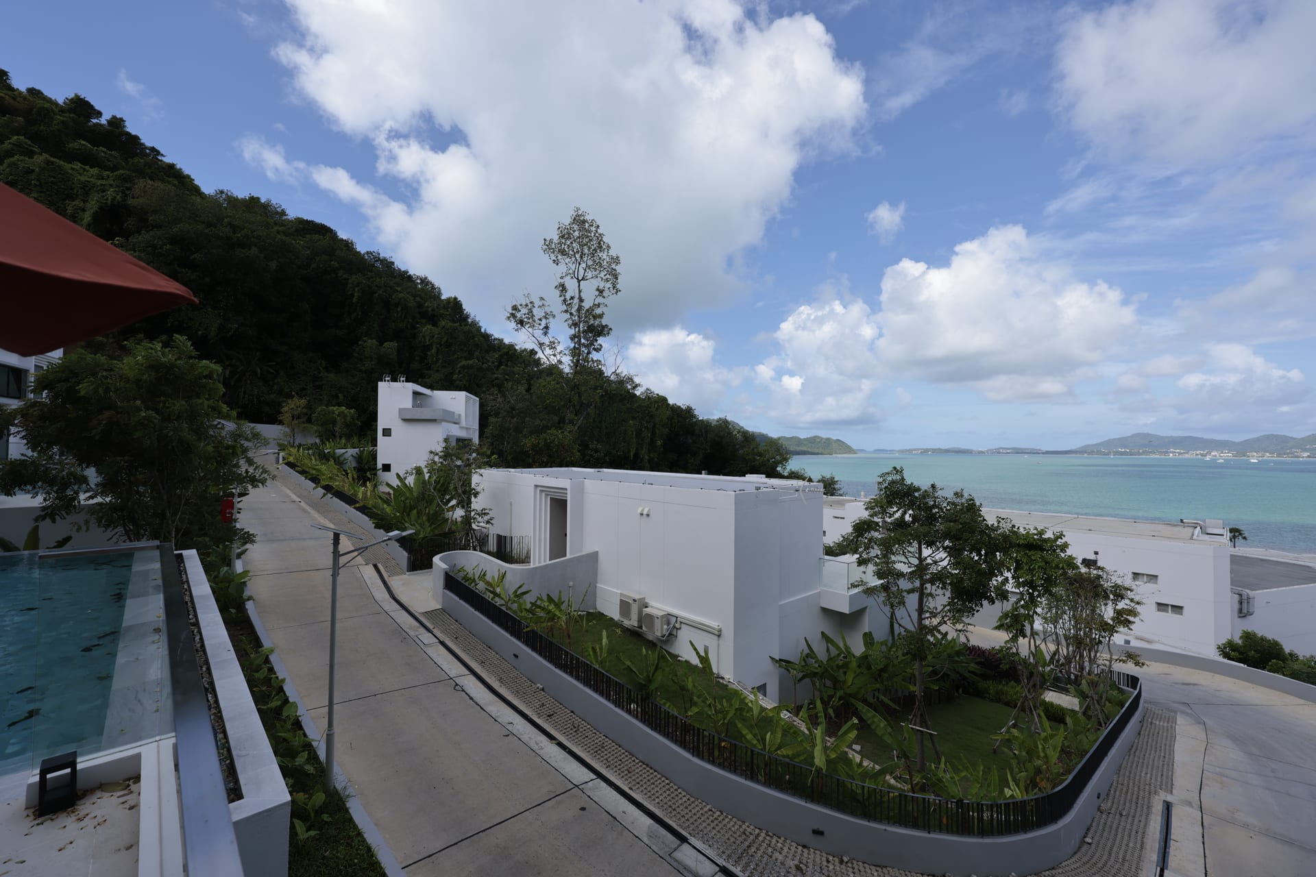 Wide view from the suite's terrace edge — the pool sits in the lower left, the pedestrian path winds down past a neighbouring villa, the bay opens up to the right with green headlands and blue water under a bright sky with scattered cumulus clouds