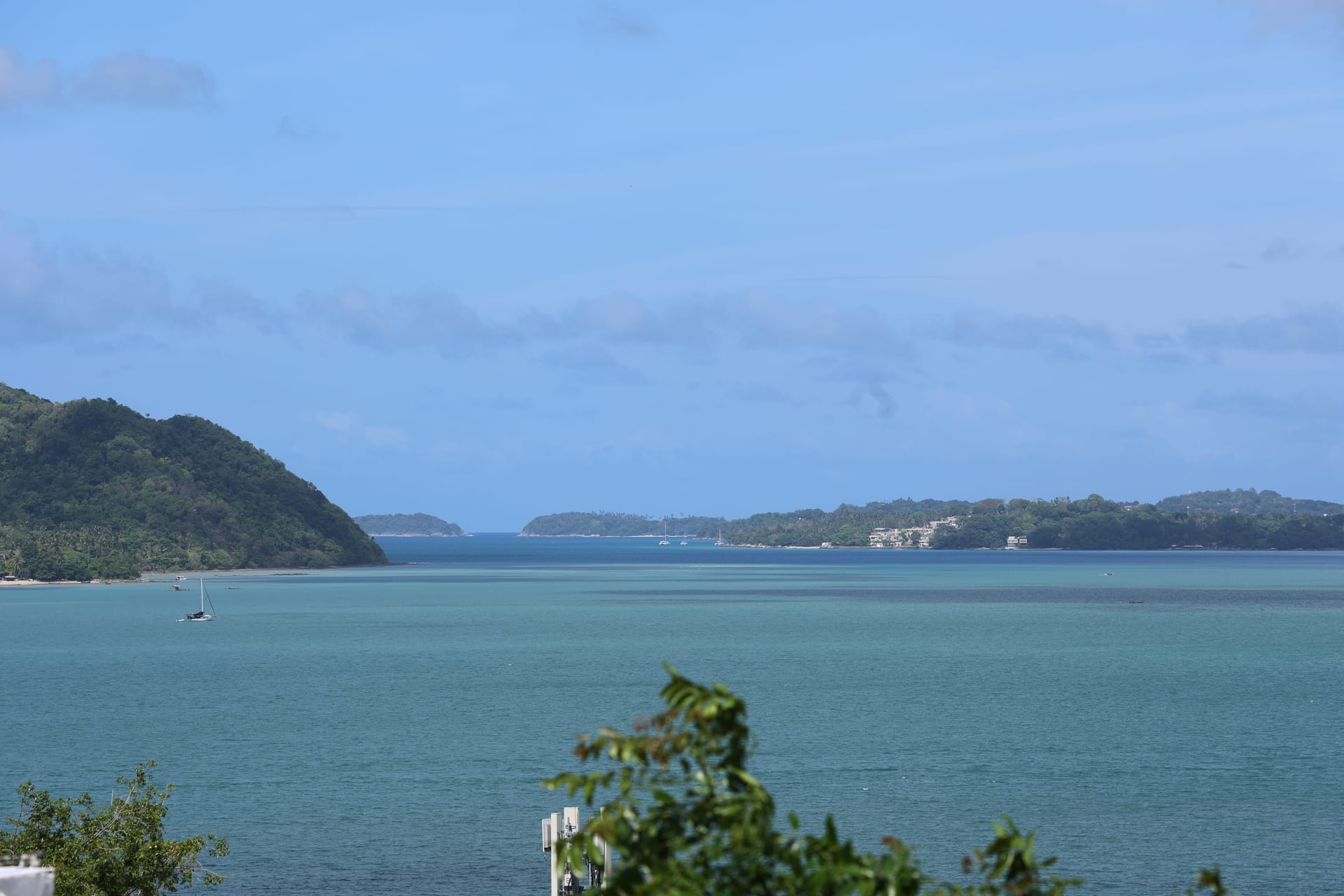 Calm bay panorama from the terrace — green headland on the left frames the channel, distant islands sit on the horizon, a lone sailboat is moored in the foreground, gentle clouds drift across a pale-blue sky