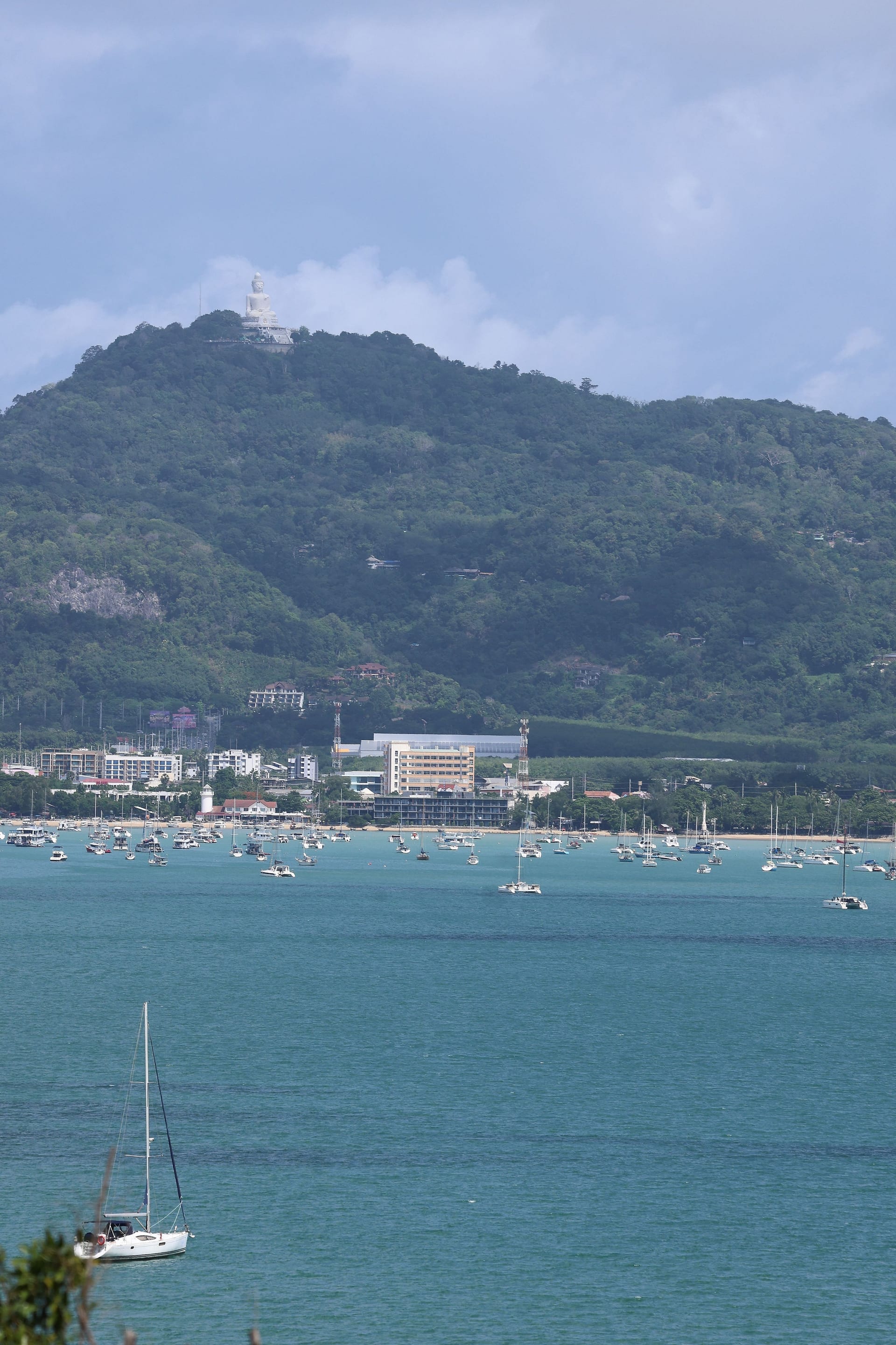Telephoto from the terrace showing Big Buddha (the 45-metre white marble statue) on the hilltop above Chalong, with Chalong town along the shoreline and the bay's sailing anchorage full of moored sailboats in the middle distance, a lone sailboat in the foreground for scale