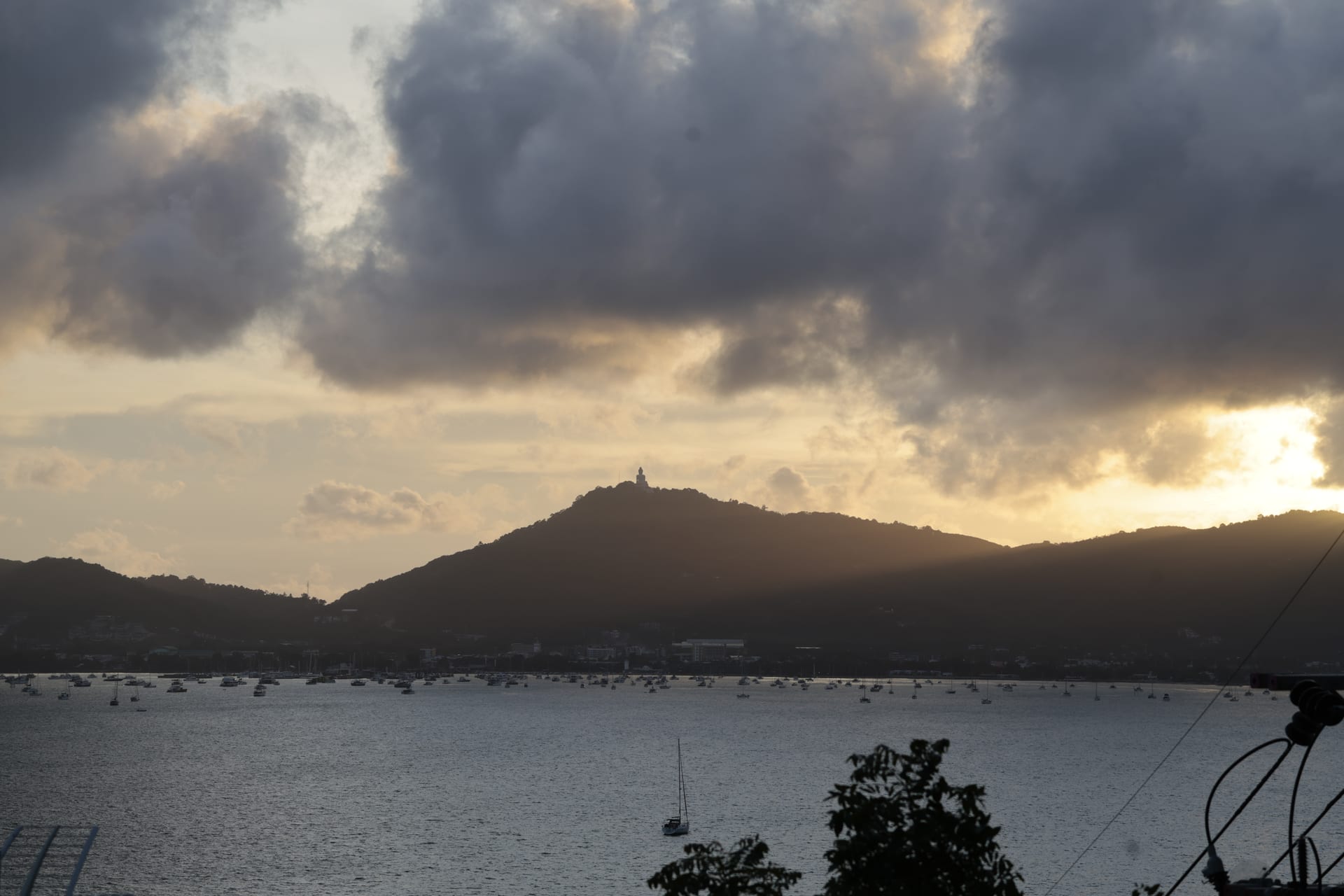 Big Buddha statue silhouetted on its hilltop above Chalong at sunset, with dramatic clouds catching the last light and the bay's moored sailboats visible in the foreground